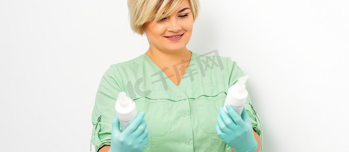 医学美容的女摄影照片_女美容师手里的化妆品和护肤品微笑着站在白墙背景上。