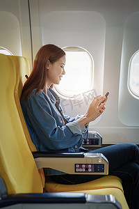 飞机飞行、旅行概念、度假时使用手机坐在经济舱靠窗座位上的亚洲女性的迷人肖像