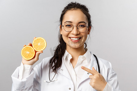 身着白大褂、戴眼镜、开朗迷人的女医生的特写镜头，指着橙色，建议吃健康食品、水果和蔬菜，以保持安全并照顾自己的健康，灰色背景