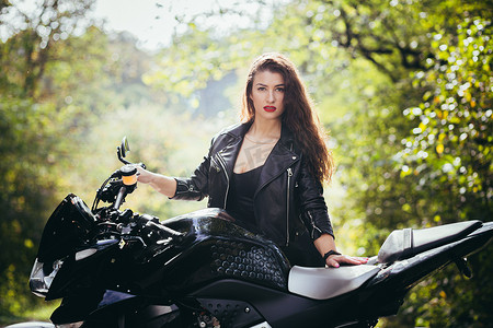 在阳光明媚的天气里，在大自然、街道、森林里，一位穿着黑色皮衣骑着摩托车的漂亮年轻女子的肖像，特写，看着相机