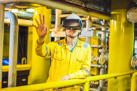身穿黄色工作服、眼镜和头盔的年轻女性在工业环境、石油平台或液化气厂使用虚拟现实眼镜