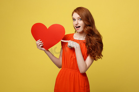 情人节和生活方式概念：一位身穿红色连衣裙的漂亮年轻女子的肖像，手指指向纸心，隔离在黄色工作室背景。