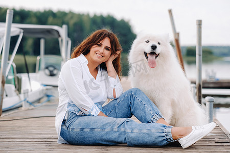 坐在海边摄影照片_日落时分，一个快乐的女人和一条大白狗坐在海边的码头上