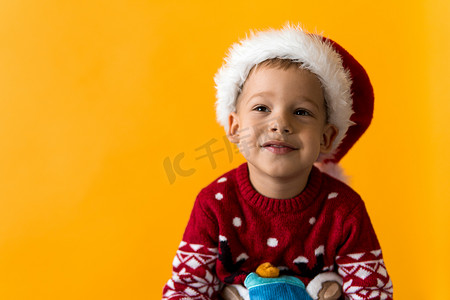 快乐微笑、积极、快乐、积极的学龄前小男孩的肖像，戴着红色温暖的圣诞老人帽子，在橙色、黄色背景上竖起大拇指。