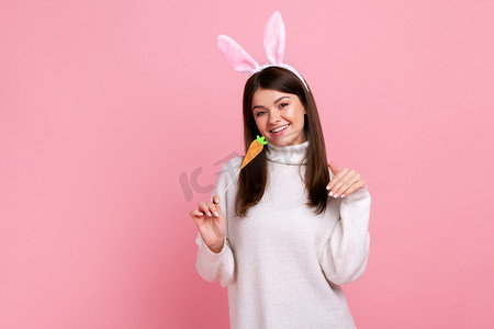 滑稽兔子摄影照片_滑稽顽皮的女孩，长着兔子耳朵，手里拿着胡萝卜站着，微笑着看着相机。