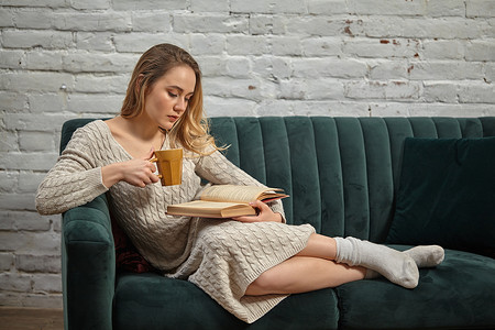 穿着米色针织连衣裙和袜子的金发模特博主斜倚在灰色沙发上，拿着棕色杯子，靠着白砖墙看书。
