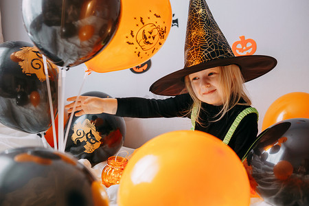 万家装饰摄影照片_儿童万圣节-一个戴着女巫帽的女孩和一个狂欢节服装，家里有通风的橙色和黑色气球。