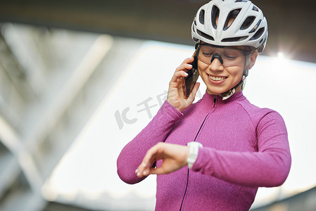 快乐的年轻女自行车手戴着防护头盔和眼镜，一边打电话一边看着手腕上的智能手表，白天站在户外
