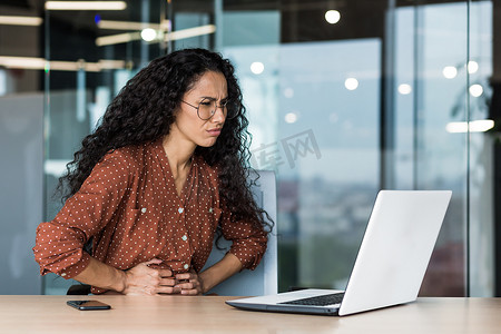 拉丁美洲女商人在工作中生病，卷发和眼镜的员工胃痛严重，女商人在办公楼内使用笔记本电脑工作。