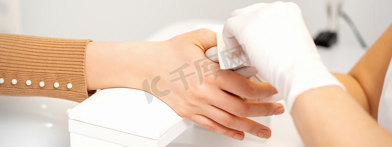 沙龙里一位戴着白色防护手套的美甲师的手用餐巾纸擦拭女性指甲。