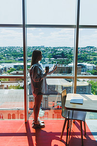 大窗户办公室摄影照片_一位戴眼镜的年轻女性自由职业者端着咖啡站在一扇大窗户旁，看着这座城市。