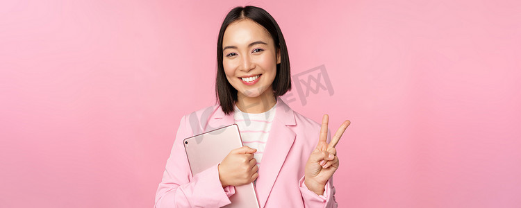 身穿西装的亚洲女商人的肖像，手拿数字平板电脑，表现出和平、V 字形和微笑、粉红色背景