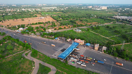 空中无人机向前飞行的镜头显示印度国道收费站的交通拥堵，汽车、卡车和车辆等待穿过周围绿树成荫的蓝色屋顶结构