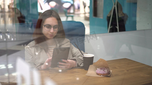 一位年轻女子坐在咖啡馆的玻璃杯后面，手里拿着一台平板电脑。