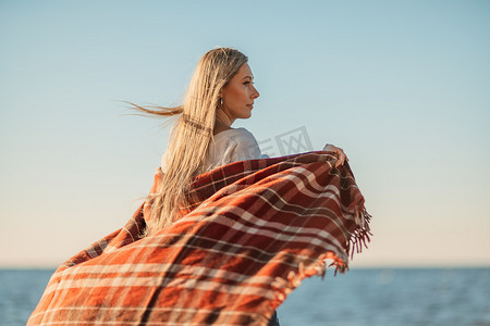 白毯子摄影照片_迷人的金发白种女人在日落时分在海滩上享受时光，裹着毯子走着，看着旁边，背景是落日的天空和大海。