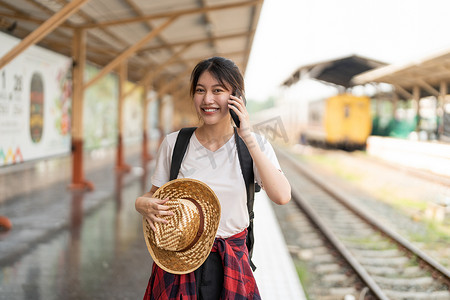 美丽的年轻亚洲女人背着背包站在站台上的火车附近使用电话。