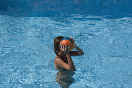 打钩图标蓝色摄影照片_晒黑的小女孩拿着球在游泳池里打水排球