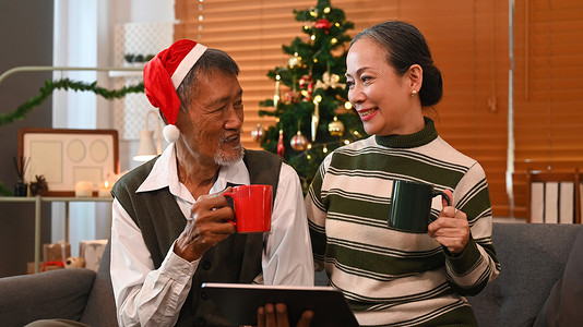 快乐的老年夫妇喜欢在装饰舒适的客厅的沙发上一起喝热巧克力并使用数字平板电脑