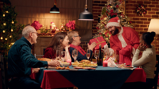 圣诞摄影照片_节日男子伪装成圣诞老人，一边享受圣诞晚餐，一边给家人送礼物