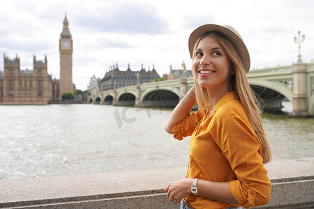 身穿橙色衬衫的自然时尚美女戴着威斯敏斯特宫和泰晤士河大桥以及英国伦敦著名大本钟塔的帽子