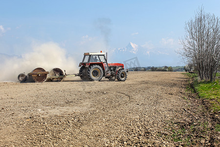 斯洛伐克 Liptovsky Hradok - 2018 年 4 月 22 日：牵引重金属滚筒的拖拉机，在春天准备干燥的田地，背后是尘云，背景是斯洛伐克标志 Krivan 山。