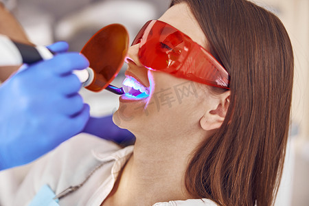 专业男医生在现代牙科柜中为快乐的白人女性补牙后使用紫外线灯