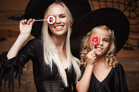 万圣节概念-美丽的白人母亲和她的女儿穿着女巫服装庆祝万圣节，在木制工作室背景上用蝙蝠和蜘蛛网庆祝万圣节糖果和糖果。