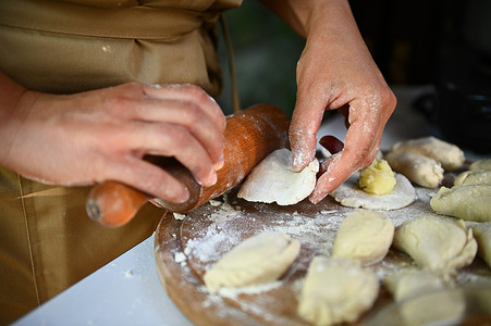 细节：穿着米色围裙的厨师的手，用 olling pin，在木板上擀面团，准备自制饺子