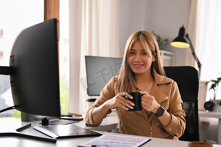 有魅力的年轻创意女性坐在家庭办公室的专业电脑前，对着镜头微笑
