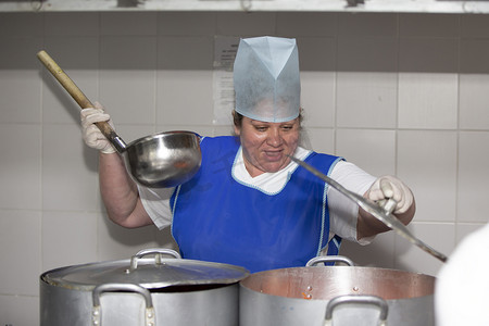学雷锋做好事摄影照片_一位女厨师在工业厨房里做饭，金属锅中放着一个大勺子。