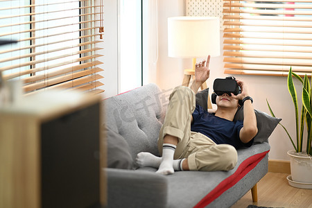 穿着休闲服的年轻人躺在沙发上看电影或在虚拟现实耳机上玩视频游戏