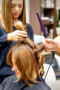 两名发型师在美容院用卷发器为顾客的棕色长发做造型。