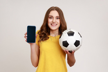 押注摄影照片_身穿黄色 T 恤、微笑积极的年轻女性手持并展示空白显示智能手机和足球，押注足球。