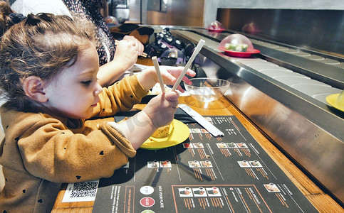 在带自助传送带的餐厅用筷子吃寿司的可爱小女孩