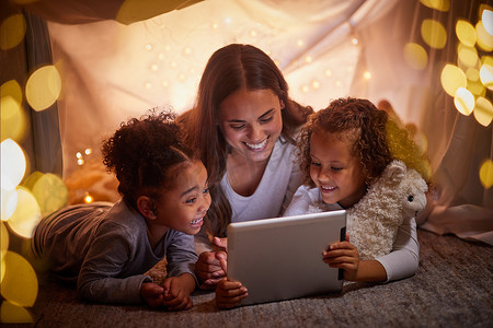 母亲和孩子们在夜间露营的帐篷里通过平板电脑或数字儿童应用程序直播流媒体电影和散景灯。