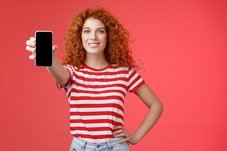 炫酷红色摄影照片_自信漂亮的红发卷发女性展示酷炫的应用程序手持智能手机扩展手显手机屏幕微笑自信推荐订阅她的博客社交媒体