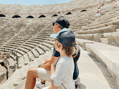圆形太阳镜摄影照片_年轻的父亲和他的小学生儿子游客在炎热的夏日参观古代古董竞技场圆形剧场遗址