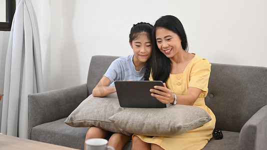 微笑的亚洲母女坐在沙发上使用数字平板电脑，享受在家一起度过的空闲时间