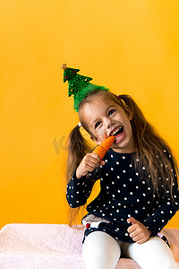 肖像正面开朗微笑快乐的小女生女孩圣诞树装饰波尔卡圆点连衣裙咬吃橙色背景上的橙色胡萝卜。