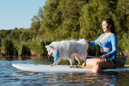 年轻女子带着她的宠物在湖上划桨，雪白的狗品种日本斯皮茨站在 Sup 板上