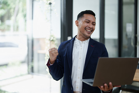 一位亚洲男性企业主站在电脑前的肖像，在成功投资后表现出幸福感
