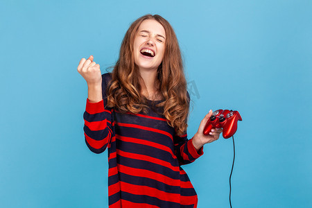 关卡游戏摄影照片_女人高兴地大喊大叫，庆祝在电子游戏中完成关卡，手里拿着红色的游戏手柄。
