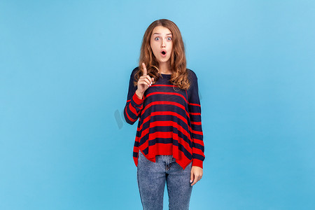 穿着条纹休闲风格毛衣的兴奋的年轻女子站起来举起手指，突然有了想法，张开嘴看着相机。