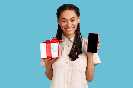 女人带着露齿的微笑看着相机，展示礼物盒和带空白屏幕的智能手机。