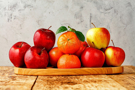 红熟的苹果和绿叶的橘子放在一张旧木桌上的木托盘上，桌子上有灰色混凝土背景。