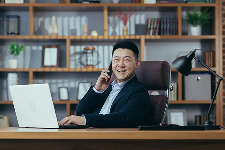 快乐的亚洲银行家老板在经典办公室里用笔记本电脑打电话