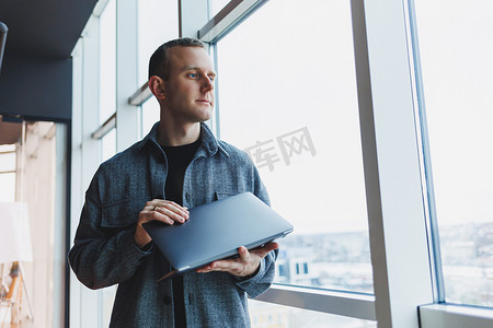 窗户台摄影照片_一个聪明聪明的男性经理的肖像，他拿着一台笔记本电脑在办公室进行研究，一个穿着优雅衣服的商人在触摸板上玩。