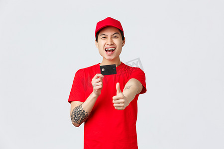 戴着红帽、T 恤制服、兴奋微笑的亚洲送货员，展示拇指和信用卡，推荐客户优惠、快递服务促销、网上购物和网上商店概念