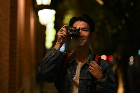 英俊的嬉皮士在城市中行走时用复古相机拍照，夜景街灯背景模糊