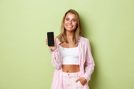 身穿粉色条纹衬衫、微笑的金发女子站在绿色背景上，展示手机屏幕的图片